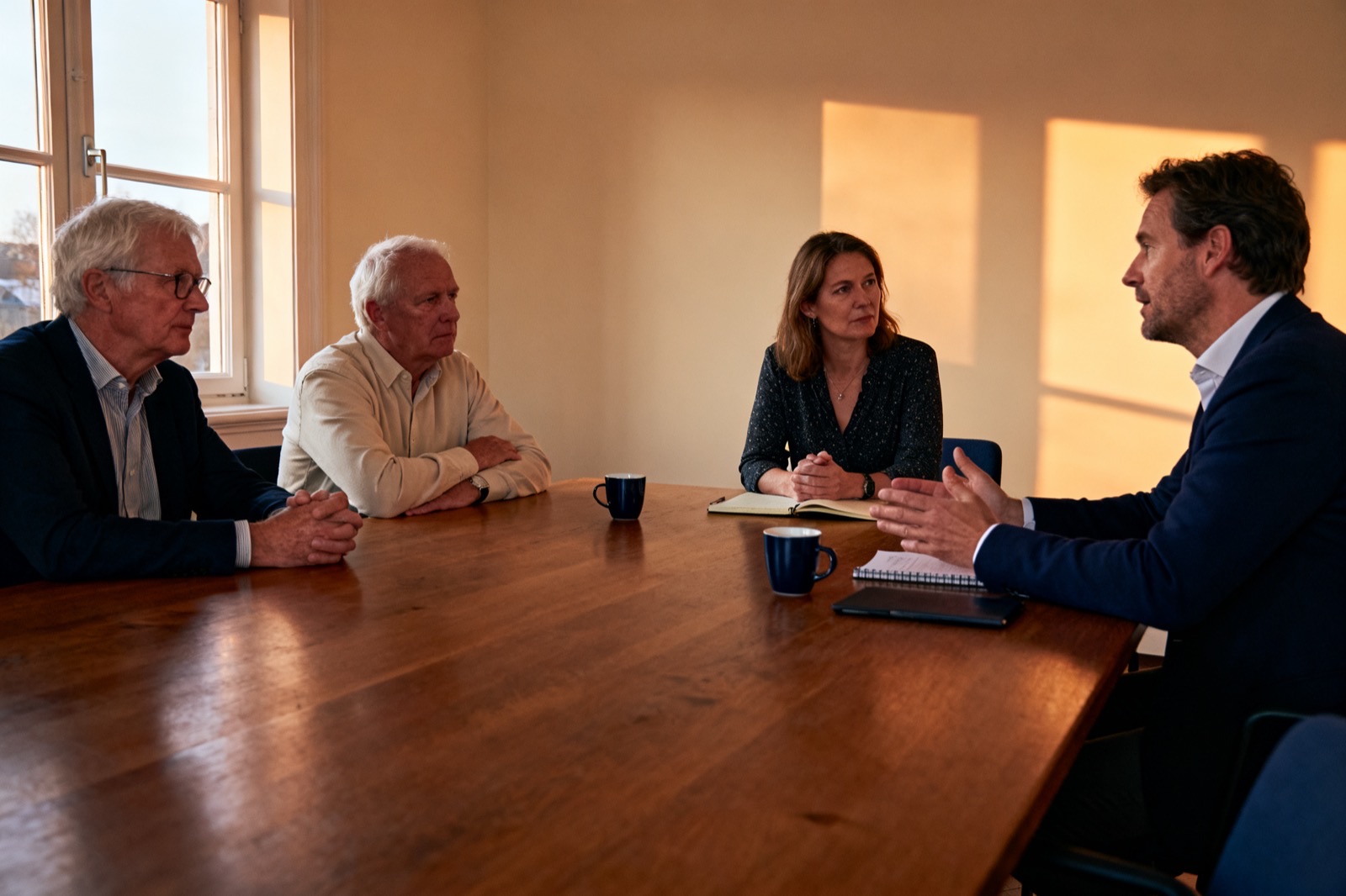 Vier mensen zitten rond een tafel in gesprek: twee ervaren mannen aan de ene kant, twee opdrachtgevers aan de andere.