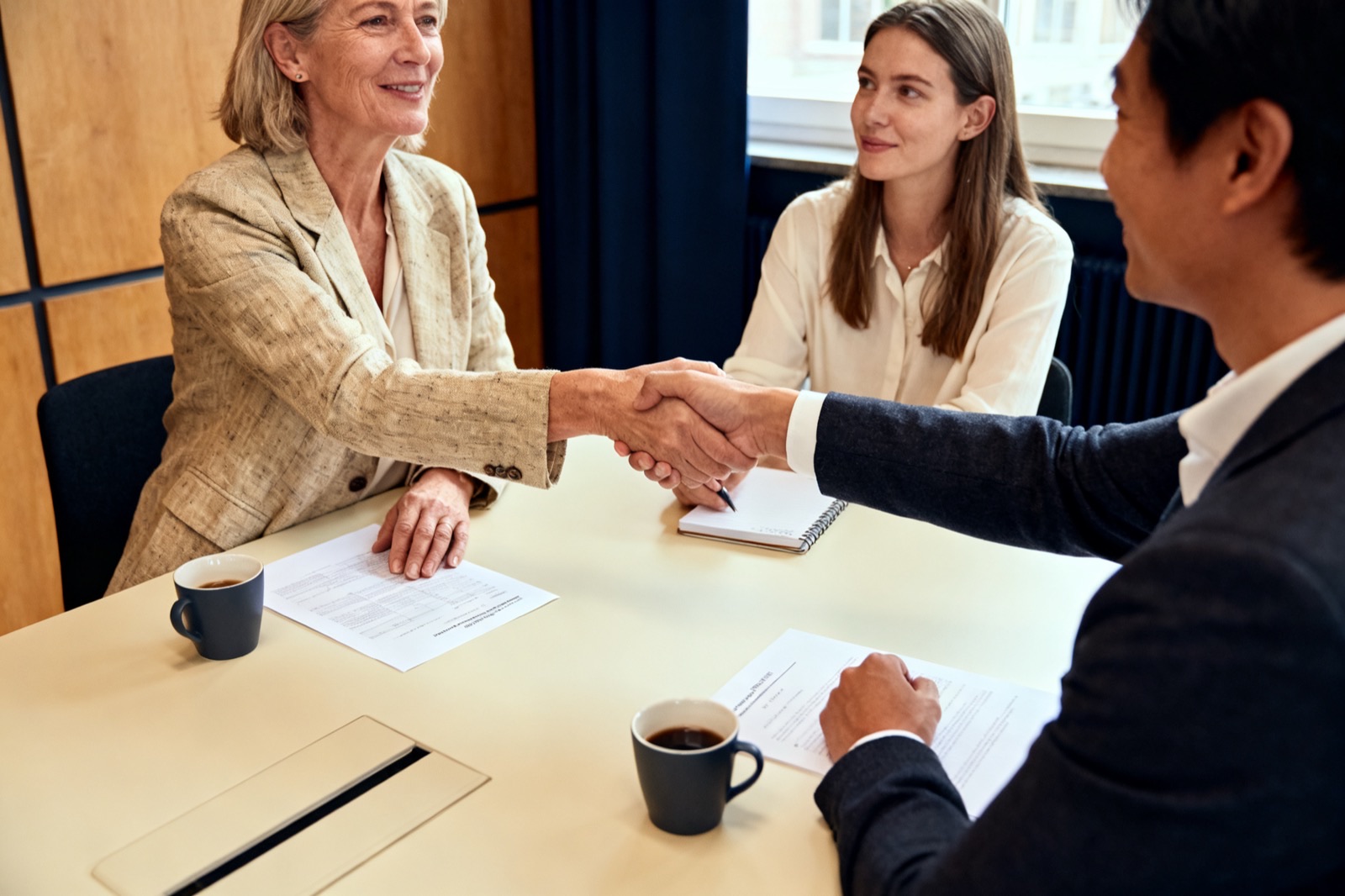 Twee partners schudden elkaar de hand aan tafel na een afspraak, terwijl een collega meekijkt.
